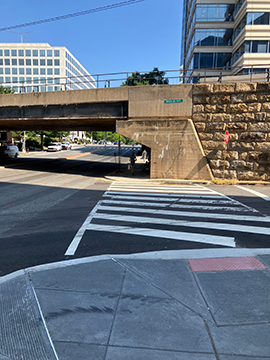 A picture of the crosswalk at Virginia Avenue that borders 7th Street. In the background there is a bridge made of concrete and stone. To the left is 7th street and to the right is Virginia Avenue. The crosswalk is on the ground past the detectable warning tile.